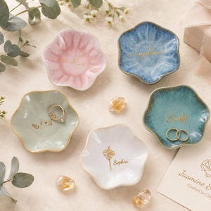May include: Five small, flower-shaped ceramic trinket dishes in pink, blue, light green, teal, and white. Each dish features gold-colored custom text or floral illustrations. Two dishes contain gold-colored rings. A branded card reads Jasmine & Sage.