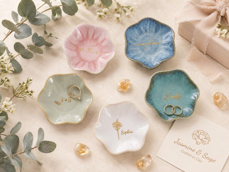 May include: Five small, flower-shaped ceramic trinket dishes in pink, blue, light green, teal, and white. Each dish features gold-colored custom text or floral illustrations. Two dishes contain gold-colored rings. A branded card reads Jasmine & Sage.
