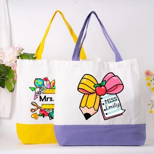 May include: Two white canvas tote bags with teacher-themed designs. One bag has a yellow base and handles, the other has a purple base and handles. The bags feature illustrations of pencils, apples, and the names "Mrs." and "Miss Emily."
