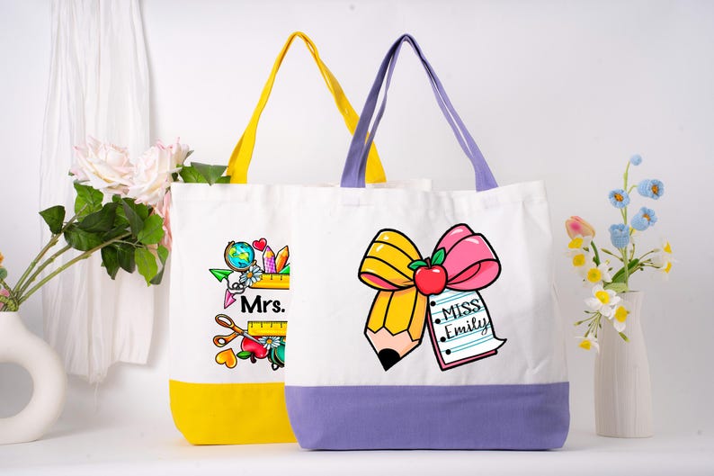 May include: Two white canvas tote bags with teacher-themed designs. One bag has a yellow base and handles, the other has a purple base and handles. The bags feature illustrations of pencils, apples, and the names "Mrs." and "Miss Emily."