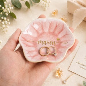 May include: A flower-shaped ceramic trinket dish with a glossy pink glaze, featuring the name Maren in gold script lettering and a small gold heart. Two gold rings rest inside the dish, which has a scalloped edge and a smooth, reflective surface.