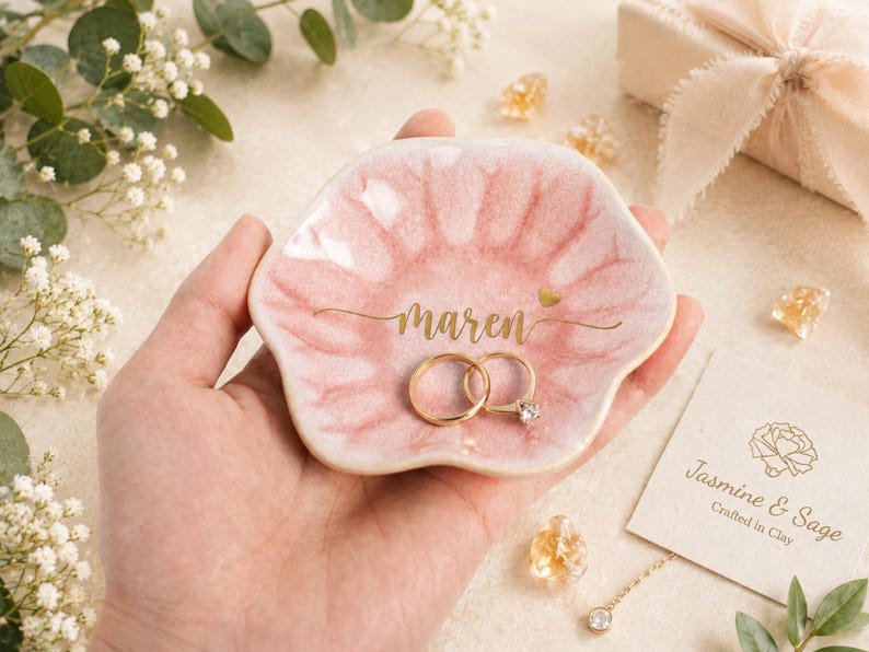 May include: A flower-shaped ceramic trinket dish with a glossy pink glaze, featuring the name Maren in gold script lettering and a small gold heart. Two gold rings rest inside the dish, which has a scalloped edge and a smooth, reflective surface.