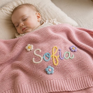 May include: A soft pink knitted baby blanket featuring the name Sofia embroidered in colourful yarn letters. The blanket is decorated with small, three-dimensional flower appliqués in white, yellow, purple, and blue, and has a ribbed border edge.