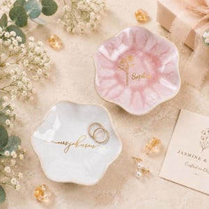 May include: Two scalloped, flower-shaped trinket dishes in soft pink and white, each featuring gold-colored script lettering. The pink dish displays a floral graphic and the name Sophia, while the white dish features cursive text and two rings.