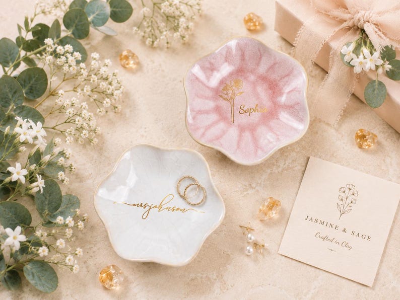 May include: Two scalloped, flower-shaped trinket dishes in soft pink and white, each featuring gold-colored script lettering. The pink dish displays a floral graphic and the name Sophia, while the white dish features cursive text and two rings.