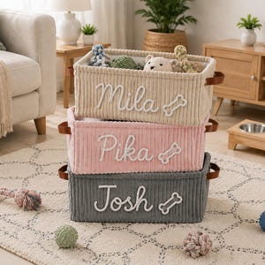 Peut inclure: Une pile de trois paniers de rangement rectangulaires en velours c&ocirc;tel&eacute; beige, rose et gris. Chaque panier poss&egrave;de des poign&eacute;es en cuir marron et des noms brod&eacute;s en blanc, Mila, Pika et Josh, avec une ic&ocirc;ne d'os brod&eacute;e sur le devant.