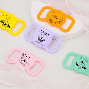 May include: Four colorful plastic dog collar buckles with personalized text and designs. The buckles are yellow, purple, pink, and green. The text on the buckles reads "Bruno", "Alain 123-341-9546", "George", and "Willow".