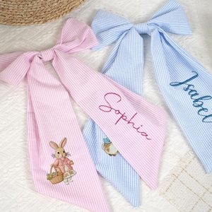 Puede incluir: Tres lazos para el pelo de tela seersucker en rosa y azul. Un lazo rosa presenta un conejito bordado con una cesta. El otro lazo rosa tiene el nombre "Sophia" bordado. El lazo azul tiene el nombre "Isabel" bordado.