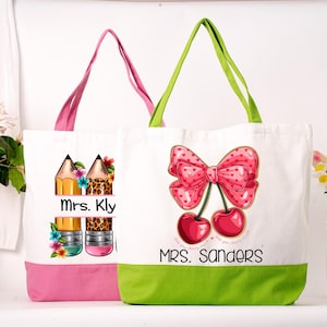May include: Two white canvas tote bags with colourful designs. One bag has a pink base and handles, featuring pencils and the text "Mrs. Kly." The other has a green base and handles, with a bow and cherries design, and the text "Mrs. Sanders."