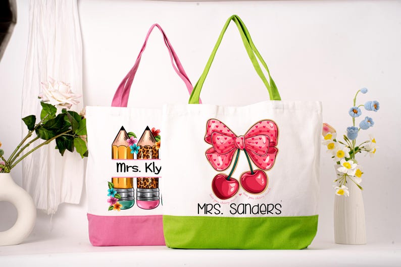 May include: Two white canvas tote bags with colourful designs. One bag has a pink base and handles, featuring pencils and the text "Mrs. Kly." The other has a green base and handles, with a bow and cherries design, and the text "Mrs. Sanders."