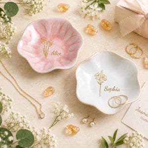 May include: Two flower-shaped ceramic trinket dishes, one in soft pink and one in white, each featuring a gold-colored floral line drawing and a name printed in script: Alice on the pink dish and Sophia on the white dish, with gold jewelry.
