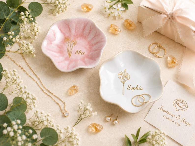 May include: Two flower-shaped ceramic trinket dishes, one in soft pink and one in white, each featuring a gold-colored floral line drawing and a name printed in script: Alice on the pink dish and Sophia on the white dish, with gold jewelry.