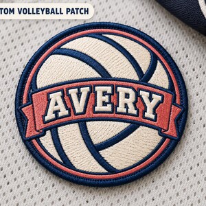 Könnte beinhalten: Ein kreisförmiger, bestickter Aufnäher mit einem cremefarbenen Volleyball mit dunkelblauen Nähten. Ein korallenfarbenes Banner verläuft durch die Mitte mit dem Namen AVERY in weißen Blockbuchstaben. Das Design hat einen dunkelblauen Rand.