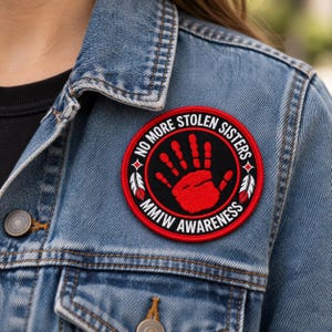 Puede incluir: Un parche bordado circular que presenta una huella de mano roja sobre un fondo negro, rodeado por el texto NO MORE STOLEN SISTERS y MMIW AWARENESS en blanco, con dos plumas blancas y rojas a los lados, fijado en una chaqueta vaquera azul.