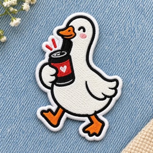 Pode incluir: Um emblema bordado apresentando um pato branco com bico e patas laranja, segurando uma lata de refrigerante preta com um rótulo vermelho e um ícone de coração branco. O pato tem uma expressão sorridente com bochechas rosas e três linhas.