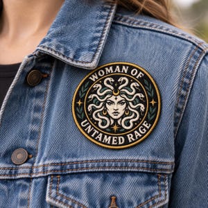Puede incluir: Un parche bordado circular que presenta una ilustración central de una figura con serpientes por cabello, rodeada por un borde negro con el texto "WOMAN OF UNTAMED RAGE" en blanco, acentuado por estrellas doradas y hojas verdes.