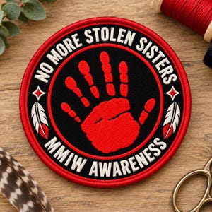 May include: A circular embroidered patch featuring a red handprint in the centre on a black background. The text NO MORE STOLEN SISTERS and MMIW AWARENESS is stitched in white around the border, accented by two white feathers with red tips.