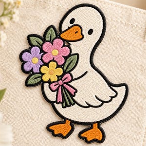 May include: An embroidered iron-on patch featuring a white duck with an orange beak and feet, holding a colourful bouquet of pink, yellow, and purple flowers with green leaves and a pink ribbon, all outlined in a thick black thread border.