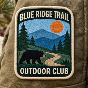 Può includere: Una toppa ricamata rettangolare con bordo color crema raffigurante un paesaggio di montagne blu, pini verdi, un sentiero beige tortuoso e un orso nero sotto un sole arancione. Il testo recita BLUE RIDGE TRAIL in alto e OUTDOOR CLUB in basso.