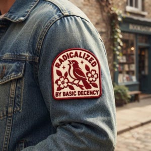 May include: A blue denim jacket featuring a rectangular embroidered patch on the sleeve. The patch has a cream background with a dark red border, a bird perched on a branch, floral accents, and the text RADICALIZED BY BASIC DECENCY.