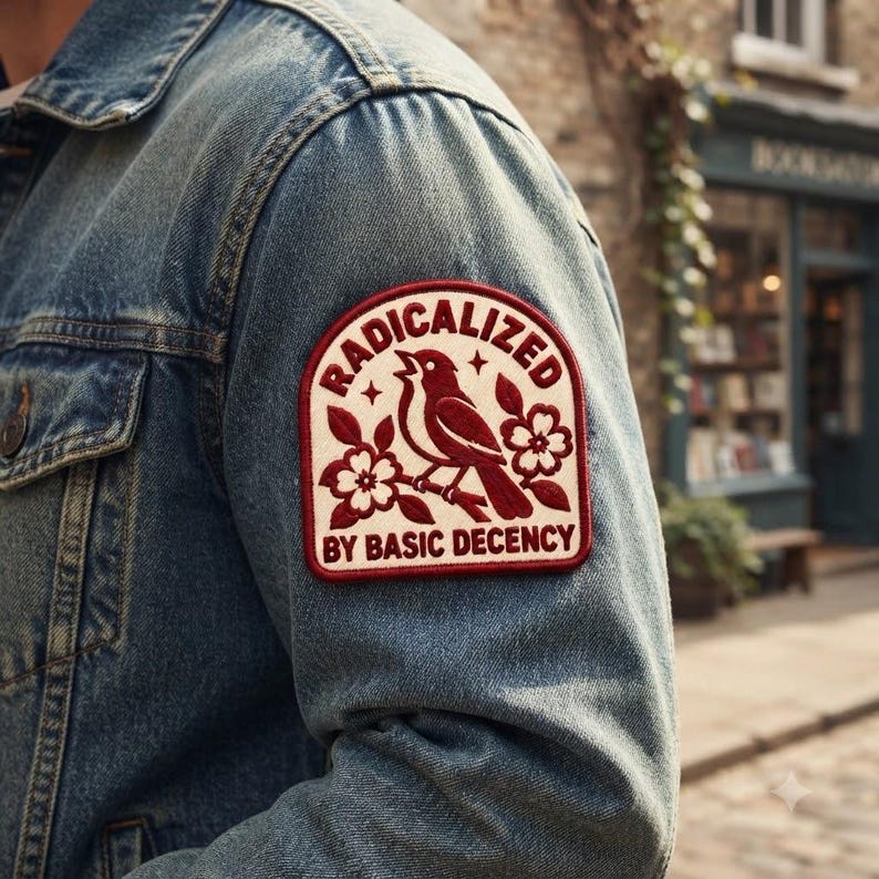 May include: A blue denim jacket featuring a rectangular embroidered patch on the sleeve. The patch has a cream background with a dark red border, a bird perched on a branch, floral accents, and the text RADICALIZED BY BASIC DECENCY.