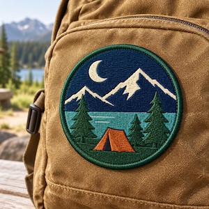 Op de afbeelding: Een ronde geborduurde patch met een nachtlandschap met een maansikkel, witte besneeuwde bergen, een blauw meer, groene dennenbomen en een oranje tent, bevestigd op het voorvak van een beige canvas rugzak.