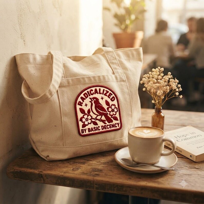 May include: A beige canvas tote bag featuring a rectangular patch with a dark red graphic of a bird perched on a flowering branch. The text RADICALIZED BY BASIC DECENCY is printed in dark red capital letters around the bird illustration.