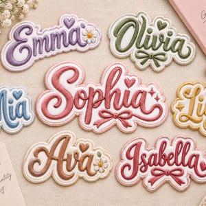 Peut inclure: Une collection d'écussons brodés avec les prénoms Emma, Olivia, Mia, Sophia, Lily, Ava et Isabella dans diverses couleurs comme le violet, vert, bleu, rose, doré, marron et rouge, ornés de cœurs, de nœuds ou de petites fleurs décoratives.
