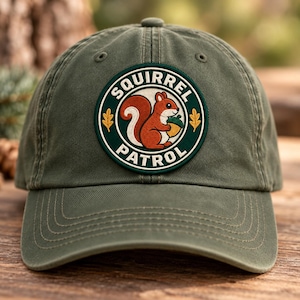 Pode incluir: Um boné de beisebol verde-oliva com um emblema circular bordado na frente. O emblema tem uma borda verde-escura com o texto SQUIRREL PATROL em branco, envolvendo uma ilustração central de um esquilo vermelho segurando uma bolota.