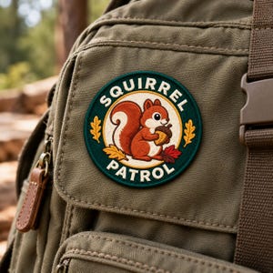 Pode incluir: Uma mochila de lona verde-oliva com um emblema circular bordado. O emblema mostra um esquilo vermelho segurando uma bolota, cercado por folhas de carvalho amarelas e vermelhas, com o texto SQUIRREL PATROL em letras maiúsculas brancas.
