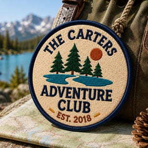 Könnte beinhalten: Ein kreisförmiger, bestickter Aufnäher mit cremefarbenem Hintergrund und dunkelblauem Rand. Er zeigt grüne Tannen, einen blauen See und eine orangefarbene Sonne. Der Text lautet THE CARTERS ADVENTURE CLUB EST. 2018 in dunkelblauer und oranger Schrift.