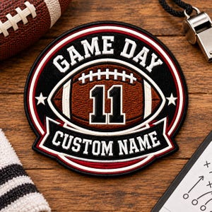 May include: A circular embroidered patch featuring a brown football graphic with the number 11 in black and white. The text GAME DAY is arched above, and CUSTOM NAME is on a banner below. The design uses black, red, and white thread colors.