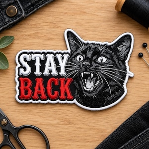 Puede incluir: Un parche bordado que muestra un gato negro con la boca abierta, mostrando los dientes, y el texto STAY BACK en letras blancas y rojas. El gato tiene bigotes blancos detallados y textura de pelaje, con un borde blanco alrededor del diseño.