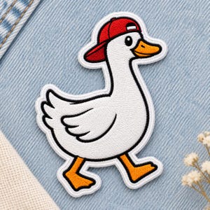 Op de afbeelding: Een geborduurde strijkpatch van een witte eend met een oranje snavel en poten, die een felrode honkbalpet draagt. De eend is afgebeeld in een zijprofiel terwijl deze loopt, met zwarte omlijningen en een witte rand op een textiel.