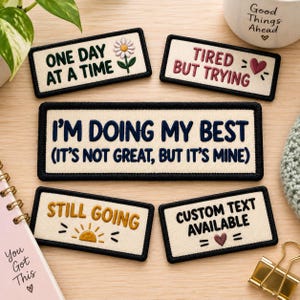 Könnte beinhalten: Fünf rechteckige bestickte Aufnäher auf cremefarbenem Stoff mit schwarzem Rand. Der Text umfasst: ONE DAY AT A TIME mit einer Blume, TIRED BUT TRYING mit einem Herz, I'M DOING MY BEST (IT'S NOT GREAT, BUT IT'S MINE), STILL GOING mit einer Sonne und CUSTOM TEXT AVAILABLE.