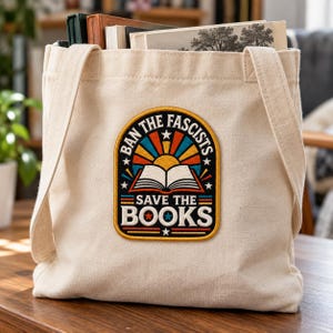 Puede incluir: Un bolso de tela beige con un parche bordado central que muestra un libro abierto, rayos de sol y el texto "BAN THE FASCISTS SAVE THE BOOKS" en colores blanco, negro, rojo, azul y amarillo con pequeñas estrellas.