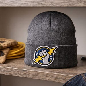 Puede incluir: Un gorro de punto gris carb&oacute;n con un parche bordado circular en el dobladillo. El parche muestra un pu&ntilde;o gris que sujeta un rayo amarillo dentado sobre un fondo azul, todo ello delineado con un grueso borde blanco.