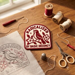 May include: A circular embroidered patch with a cream background and dark red border featuring a bird perched on a branch with flowers and the text RADICALIZED BY BASIC DECENCY. A matching outline is printed on a piece of fabric nearby.