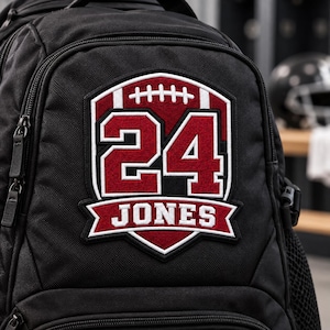 Puede incluir: Una mochila negra con un gran parche bordado en la parte delantera. El parche tiene forma de escudo en color granate y blanco, con un gráfico de balón de fútbol, el número 24 en letras grandes y el nombre JONES en blanco.