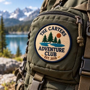 May include: An olive green backpack featuring a circular embroidered patch with a tan background. The patch displays a blue lake, green pine trees, an orange sun, and the text THE CARTERS ADVENTURE CLUB EST. 2018 in dark blue lettering.