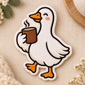 Pode incluir: Um emblema bordado apresentando um ganso branco com contorno preto, segurando uma caneca castanha com vapor a sair. O ganso tem pés e bico cor de laranja, com um pequeno rubor rosa na bochecha e um olho fechado a sorrir.