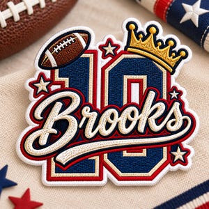 Puede incluir: Un parche bordado que muestra el número 10 en azul marino con bordes rojos y crema. El diseño incluye un balón de fútbol americano marrón, una corona dorada y varias estrellas color crema. El nombre Brooks está bordado en letra cursiva crema.