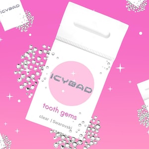 May include: White packets of tooth gems with the brand name "ICYBAD" in glittery silver letters on a pink circle. The text "tooth gems" and "clear | Swarovski" are below the circle. The background is pink with scattered gems.