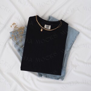 Op de afbeelding: Een zwart shirt met een mock neck, gevouwen op een paar lichtblauwe jeans. Een gouden ketting met een hanger ligt over het shirt.