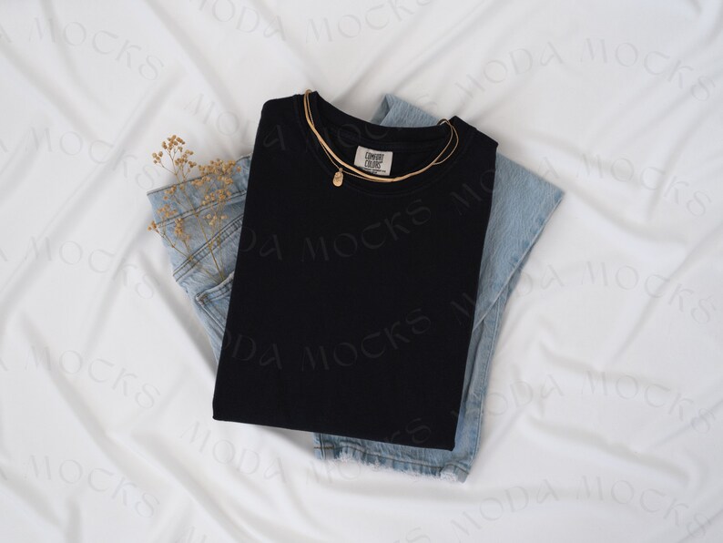 Op de afbeelding: Een zwart shirt met een mock neck, gevouwen op een paar lichtblauwe jeans. Een gouden ketting met een hanger ligt over het shirt.