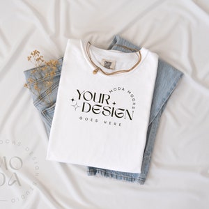 May include: A white t-shirt with the text "YOUR DESIGN GOES HERE" is folded on top of a pair of light blue jeans. A gold necklace and dried flowers are also present. The items are arranged on a white fabric background.