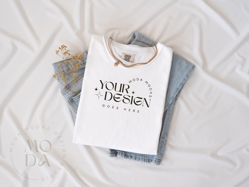 May include: A white t-shirt with the text "YOUR DESIGN GOES HERE" is folded on top of a pair of light blue jeans. A gold necklace and dried flowers are also present. The items are arranged on a white fabric background.