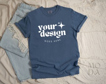 May include: A blue t-shirt with a white graphic that says "your design goes here" with a star. The shirt is laying on a white and beige blanket with a pair of blue jeans.