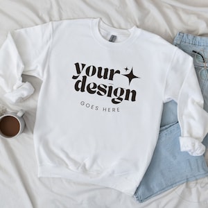 May include: White sweatshirt with a black graphic that says "your design goes here" with a star on the right side of the text. The sweatshirt is laid flat on a white bedspread with a pair of blue jeans and a coffee mug.