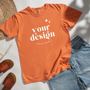 May include: A bright orange t-shirt with the text "your design goes here" in white. The shirt is laid flat on a white surface with a straw hat, sunglasses, and denim shorts.